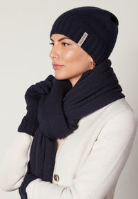 Marinblå stickad mössa med vikbar ribbad kant, matchande scarf med mjuk textur och en diskret logotyp. Stylad med en lätt cardigan.