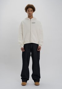 Felpa cremosa con zip, dotata di tasca frontale e logo ricamato, abbinata a pantaloni di jeans scuri larghi e stivali color camoscio. Texture liscia, vestibilità casual.