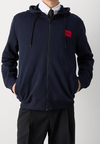 Hombre con sudadera con cremallera color navy y cordones negros, parche rojo "HUGO", camisa de cuello blanca, corbata negra y pantalones negros.