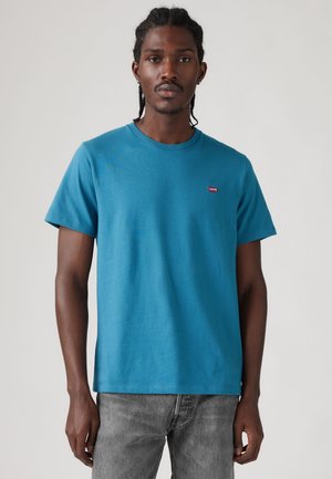 Jeune homme aux cheveux tressés portant un t-shirt Levi's bleu sarcelle et un jean gris, debout devant un fond clair et uni.