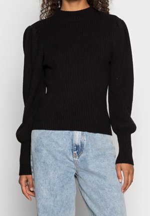 Vrouw die een zwarte geribbelde trui met opgeblazen lange mouwen en lichtblauwe high-waisted jeans draagt, staand tegen een neutrale achtergrond.