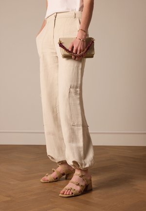 Beige linnen broek met een relaxed fit en zakken, gecombineerd met tan gekleurde sandalen met hakken en gespen. Een handtas in beige met een kettingriem.
