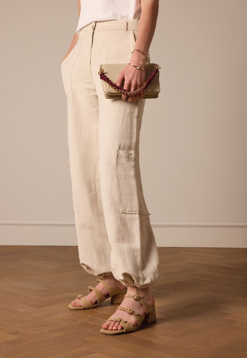 Beige linnen broek met een relaxed fit en zakken, gecombineerd met tan gekleurde sandalen met hakken en gespen. Een handtas in beige met een kettingriem.