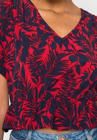 Rote Blumenbluse mit V-Ausschnitt, kurzen Ärmeln, geraffter Taille und marineblauen Blattmustern aus einem weichen, leichten Stoff.