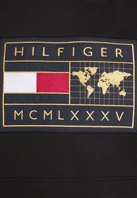 Tessuto nero con un patch ricamato in oro con "HILFIGER", una mappa e strisce orizzontali rosse e bianche. Inclusa l'anno "MCMLXXXV".