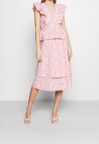 Vaaleanpunainen kukkamekko, jossa on röyhelöhihat, v-kaula-aukko, vartalonmyötäinen yläosa ja kerroksittainen hame. Yhdistetty valkoisiin remmikenkiin.