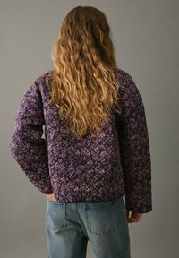Giacca trapuntata viola con motivi floreali, maniche lunghe e vestibilità rilassata. Abbinata a jeans blu, vista da dietro su uno sfondo neutro.