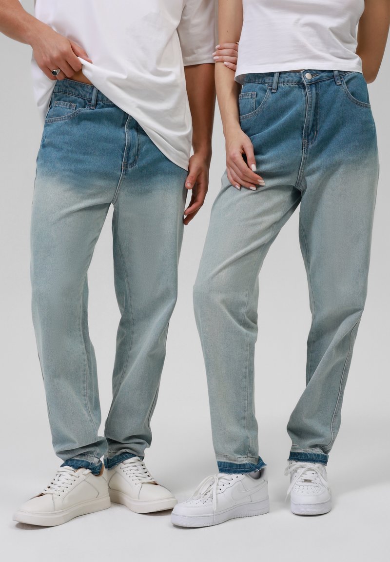 Deux personnes portant des jeans bleus ombrés, des baskets blanches et des t-shirts blancs, debout côte à côte, l'une tenant le bras de l'autre.