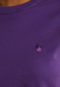 Camiseta de manga corta púrpura, hecha de algodón suave, con un pequeño logo bordado en el lado izquierdo del pecho. Textura suave y corte clásico.