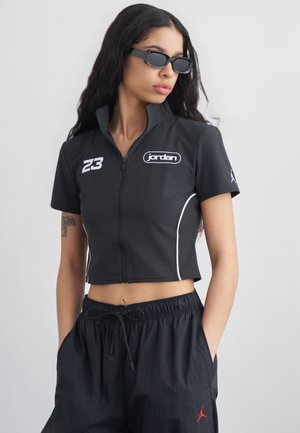 Frau trägt eine schwarze kurze Jacke mit „jordan“ Logo und der Nummer 23, schwarze Hose mit rotem Logo und rechteckige schwarze Sonnenbrille.