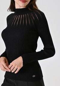Maglione nero a coste con scollatura alta e dettaglio in pannello trasparente. Presenta maniche lunghe e un piccolo logo sul bordo.