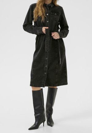 Kvinde iført en lang sort denim kjole med knapper og bælte i taljen samt knæhøje sorte læderstøvler med spids snude og hæl.