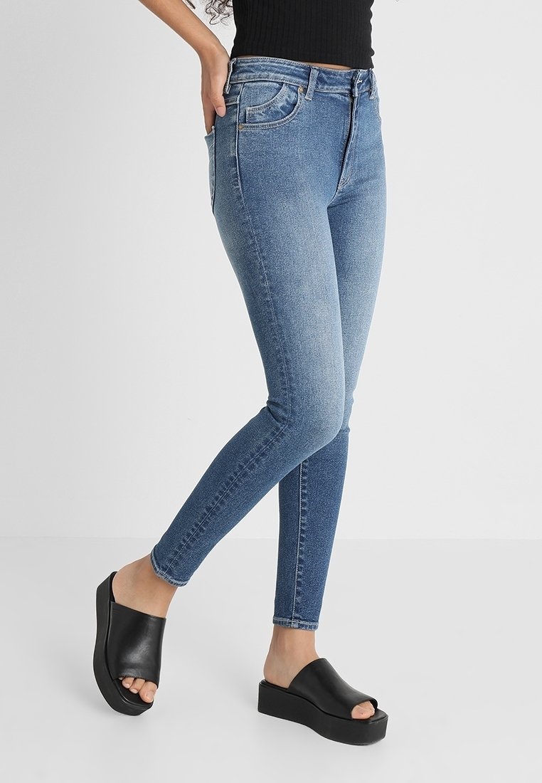 Jeans ajustados de mezclilla azul medio, con cintura alta, un sutil desgastado y diseño de cinco bolsillos. Combinados con sandalias de plataforma negras.