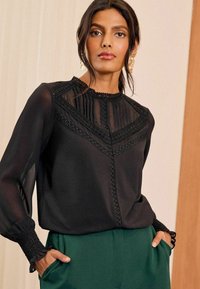 Schwarze Bluse mit hohem Kragen, plissierter Vorderseite und aufwendiger Spitzenverzierung. Lange Ärmel mit gerafften Manschetten. Kombiniert mit grünen Hosen.