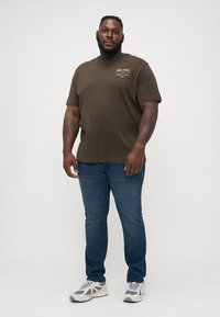 Brun bomulds t-shirt med korte ærmer og et lille trykt tekstdesign, parret med blå jeans og grå sneakers.