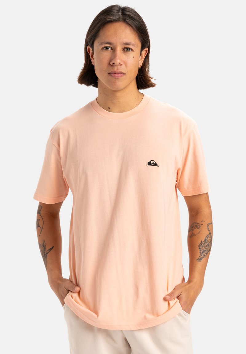 Camiseta de algodón de color rosa claro con cuello redondo, mangas cortas y un pequeño logo negro en el pecho izquierdo. Textura suave.