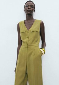 Olive green jumpsuit with a V-neck, two front pockets, and button closure. Features tailored design and wide-leg pants.