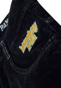 Crne korduroy hlače s izvezenim "homeboy x-tra" logotipom u narančastoj i tirkiznoj boji na džepu, s vidljivim detaljima šavova.