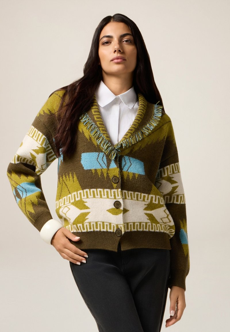 Neulecardigan oliivinvihreänä, jossa on geometristä kuviota valkoisena ja sinisenä. Varustettu huivikauluksella, fringe-yksityiskohdilla ja nappikiinnityksellä.