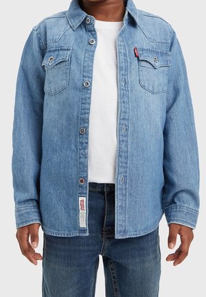 Personne portant une veste en jean bleu clair sur une chemise blanche et un jean bleu foncé, debout avec les bras détendus le long du corps.