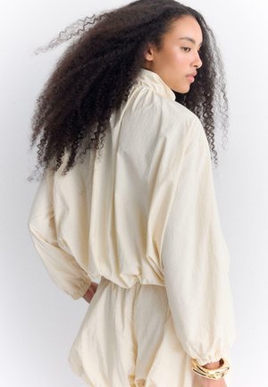 Žena s dugom kovrčavom kosom u širokoj krem bluzu i odgovarajućim kratkim hlačama okreće glavu u stranu na bijeloj pozadini.