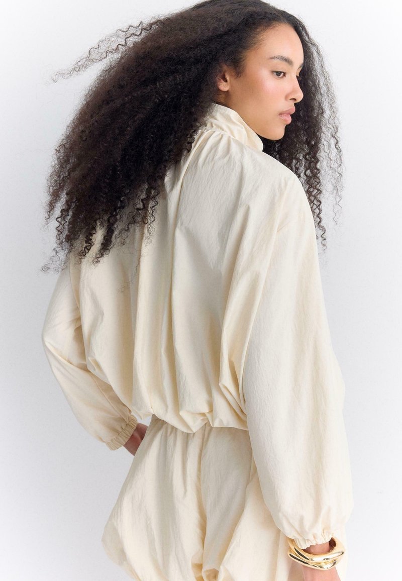 Femme aux longs cheveux bouclés portant un chemisier ample crème et un short assorti, tournant la tête sur le côté sur fond blanc.