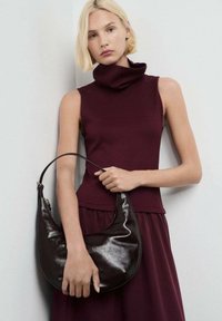 Una mujer con cabello corto y rubio lleva puesto un vestido de cuello alto sin mangas de color burdeos, sosteniendo un gran bolso de piel de cuero marrón oscuro brillante.