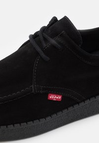 Černé semišové volnočasové šněrovací boty s tlustou texturovanou podrážkou a malou červenou textilní etiketou na vnější straně poblíž švu.