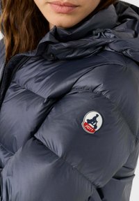 Marineblauwe pufferjack met een doorgestikt ontwerp, glanzende afwerking en logopatch op de mouw. Heeft een capuchon en een veilige ritssluiting.