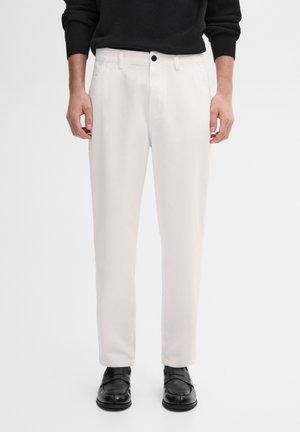 Homme portant un pantalon blanc droit, des mocassins noirs et un haut noir à manches longues, debout devant un fond uni.