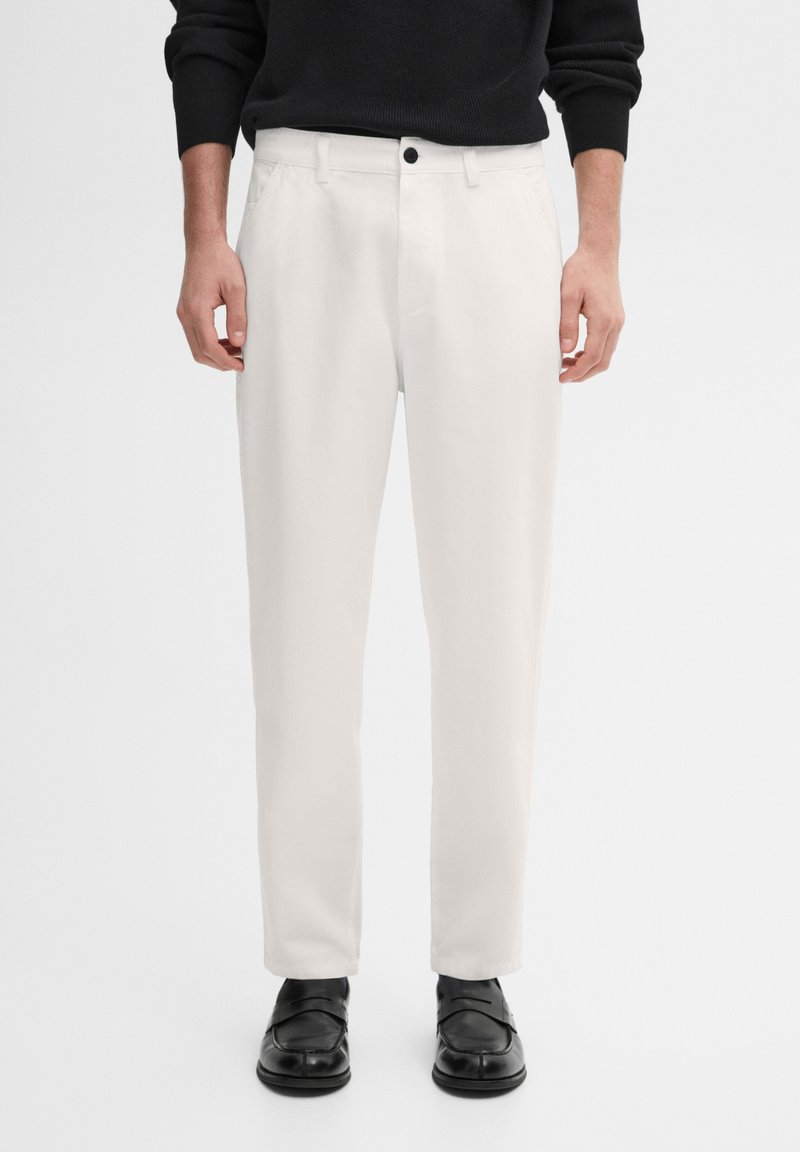Homme portant un pantalon blanc droit, des mocassins noirs et un haut noir à manches longues, debout devant un fond uni.