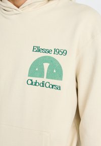 Hanorac bej cu buzunar frontal, având un logo brodat verde: "Ellesse 1959" și "Club di Corsa" cu un design geometric. Textură moale.