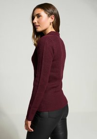 Frau in einem gerippten burgunderfarbenen Pullover und schwarzen Hosen, die seitlich vor einem schlichten hellen Hintergrund steht.