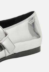 Silver patent leather shoe with a low heel, featuring a strap and rectangular buckle detail. Smooth texture with visible stitching.