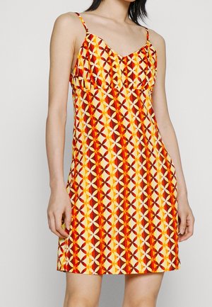 Vrouw die een mouwloze jurk draagt met een rood, geel en wit geometrisch patroon en dunne schouderbandjes, staand tegen een effen achtergrond.