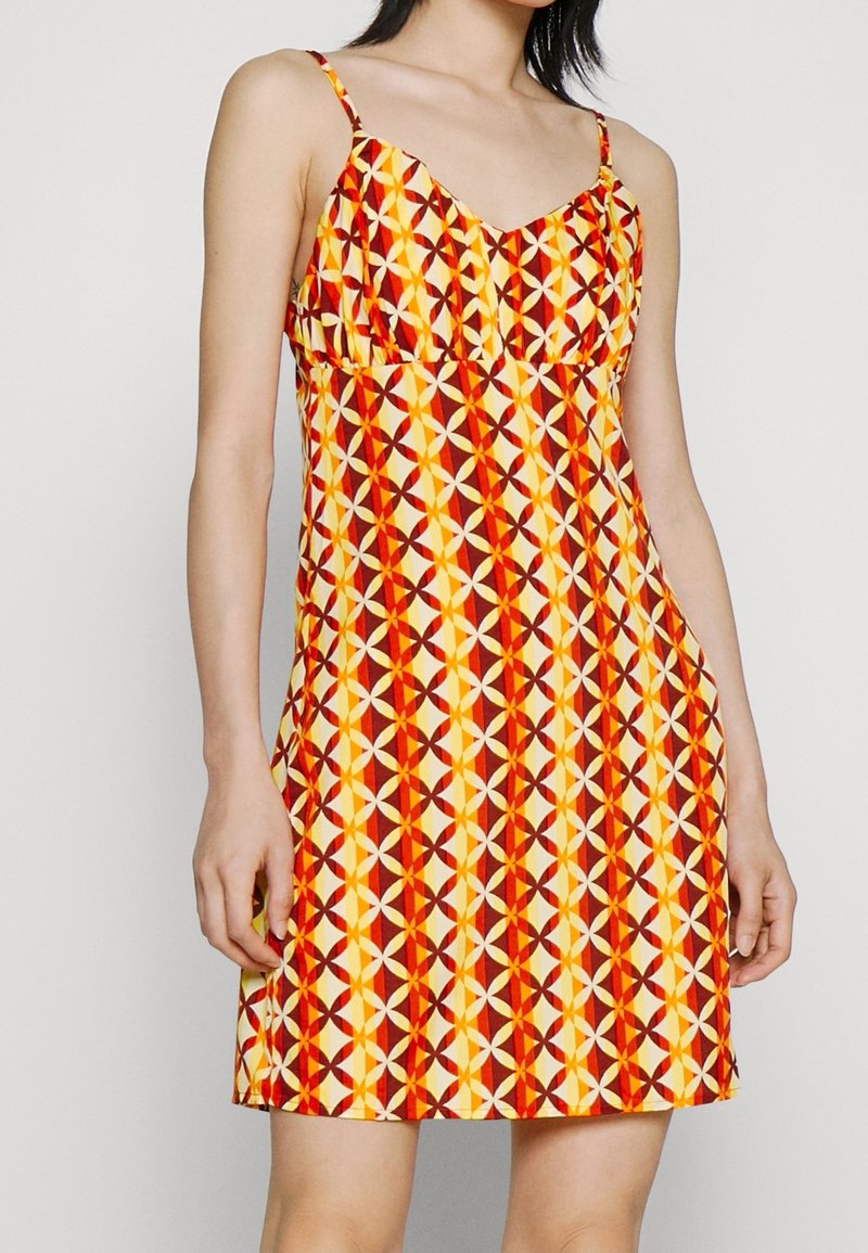 Femme portant une robe sans manches avec un motif géométrique rouge, jaune et blanc, avec de fines bretelles, debout devant un fond uni.