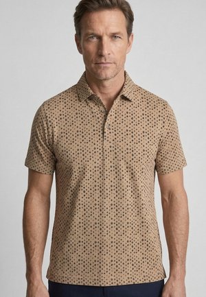 Man wearing a tan short-sleeve polo shirt with a small black circular pattern, standing against a plain light background.