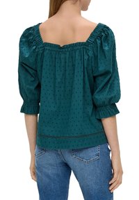 Kobieta ubrana w teksturowaną bluzkę w kolorze teal z bufiastymi rękawami o długości trzy czwarte oraz falbaną elastyczną dekoltą, w połączeniu z niebieskimi dżinsami, pokazana od tyłu.