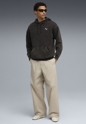 Young man wearing black Puma hoodie, beige wide-leg pants, white sneakers, and black sunglasses standing with hands in pockets.