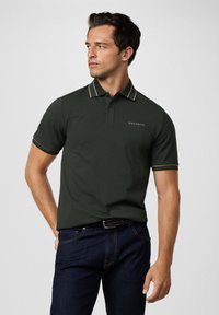 Man in een donkergroen polo shirt met beige details en het "Hackett" logo, gecombineerd met donkerblauwe jeans en een gevlochten riem, kijkend naar links.