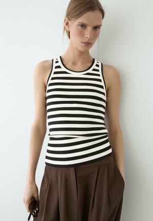 Femme portant un débardeur rayé noir et blanc et un pantalon marron ample, debout devant un fond uni clair.