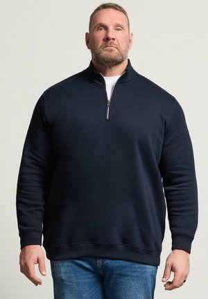 Uomo barbuto che indossa un pullover blu navy con zip a quarto sopra una camicia bianca e jeans blu, in piedi di fronte a uno sfondo chiaro uniforme.
