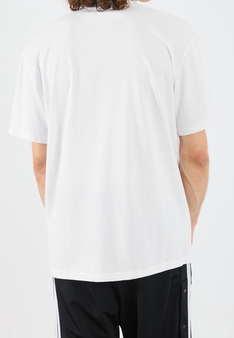 Homme aux cheveux bouclés portant un t-shirt blanc uni à manches courtes et un pantalon de survêtement noir, vu de dos sur un fond blanc.