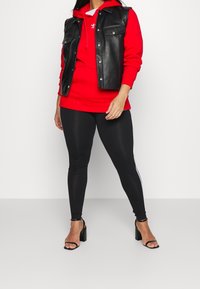 Svart läder utan ärmar jacka över en röd huvtröja, tillsammans med svarta leggings med vita sidostripes och svarta klacksandaler.