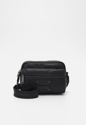 Czarna torebka na ramię Valentino z przednią kieszenią na suwak, skórzaną teksturą oraz regulowanym, materiałowym paskiem z logo na białym tle.