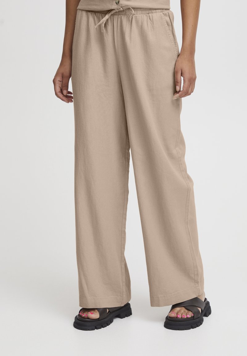 Pantalon beige à jambes larges avec taille élastique et cordon, porté avec des sandales noires épaisses montrant des ongles de pieds vernis.