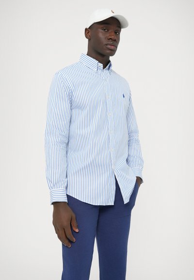 Plava i bijela prugasta košulja s gumbima i dugim rukavima, s ovratnikom i malim logotipom na prsima, u kombinaciji s tamnoplavim hlačama.
