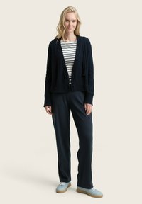 Navy cardigan with cable knit design over a white and navy striped shirt, paired with dark blue trousers and light blue sneakers.