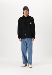 Zwarte overhemd met knopen en een zak en logo patch, gecombineerd met losse blauwe spijkerbroeken en witte sneakers met rode strepen, gedragen met een pet.
