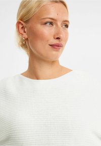 Biała ribanowa bluzka z szerokim dekoltem, ozdobiona teksturowym wzorem. Złote kolczyki w kształcie kółek dopełniają stylizację. Kolor skóry wygląda naturalnie.